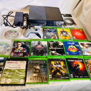 May include: A black Xbox One console with a power brick, a grey wireless controller, and a collection of seventeen video games including FIFA 14, FIFA 18, Madden 17, Starlink, Constructor, Mortal Kombat X, Fallout 76, Overwatch, Injustice 2, and Call of Duty.
