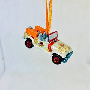 May include: A white and red toy jeep with orange accents and black wheels, suspended by an orange ribbon. The jeep features the Jurassic Park logo and a weathered appearance, suggesting a vintage or themed design.