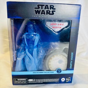 May include: A Star Wars Black Series Holocomm Collection Darth Vader action figure in a blue and black box. The figure is translucent blue and comes with a lightsaber. The box has the Star Wars logo and a warning about small parts.