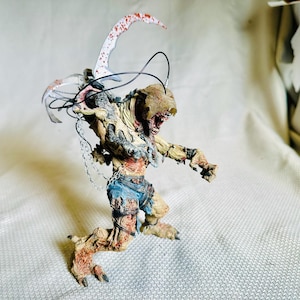 Puede incluir: Figura detallada de una criatura parecida a un zombi con una gran cuchilla curva. La figura tiene carne expuesta, ropa hecha jirones y una cadena. La cuchilla tiene salpicaduras rojas, lo que sugiere un tema violento.