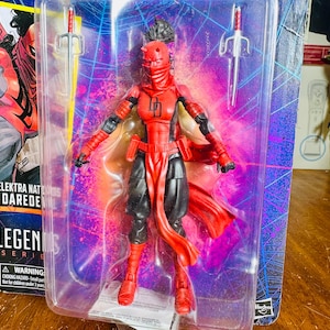 K&ouml;nnte beinhalten: Elektra Natchios Daredevil Actionfigur in einer transparenten Kunststoffverpackung. Die Figur ist &uuml;berwiegend rot und schwarz, mit zwei silbernen Sai-Waffen. Die Verpackung tr&auml;gt das Legends Series-Logo und einen Warnhinweis zu Erstickungsgefahren.