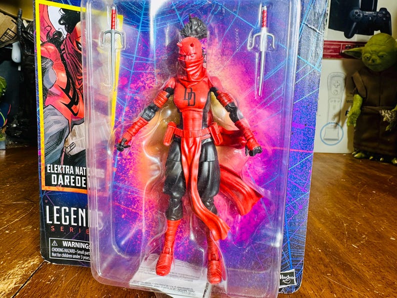 K&ouml;nnte beinhalten: Elektra Natchios Daredevil Actionfigur in einer transparenten Kunststoffverpackung. Die Figur ist &uuml;berwiegend rot und schwarz, mit zwei silbernen Sai-Waffen. Die Verpackung tr&auml;gt das Legends Series-Logo und einen Warnhinweis zu Erstickungsgefahren.