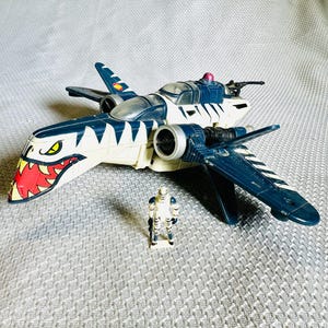 May include: A toy airplane with a shark-like design, featuring a white body with blue accents and a red and yellow mouth. A small action figure stands nearby. The toy has wings and a cockpit.
