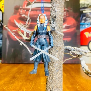 May include: A blue and white Star Wars character ornament with blue lightsabers. The ornament is hanging from a tree branch.