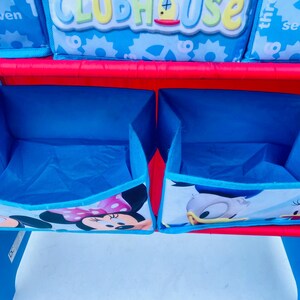 Puede incluir: Un mueble de almacenamiento azul y rojo para ni&ntilde;os con dos cubos abiertos. Los cubos est&aacute;n decorados con im&aacute;genes de Mickey y Minnie Mouse, y Donald y Daisy Duck. La estanter&iacute;a superior tiene cajas de almacenamiento con la palabra "Clubhouse".