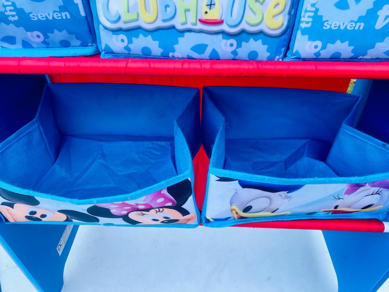 Puede incluir: Un mueble de almacenamiento azul y rojo para ni&ntilde;os con dos cubos abiertos. Los cubos est&aacute;n decorados con im&aacute;genes de Mickey y Minnie Mouse, y Donald y Daisy Duck. La estanter&iacute;a superior tiene cajas de almacenamiento con la palabra "Clubhouse".
