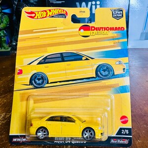 May include: A yellow Hot Wheels die-cast car in its original packaging. The packaging features a yellow and blue design with the text "Deutschland Design" and an illustration of a yellow Audi S4 quattro. The car is a 1:64 scale model.