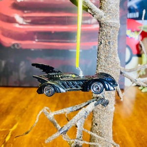 May include: A black die-cast toy car shaped like the Batmobile hanging from a branch of a small artificial tree. The car has a yellow ribbon attached to it.