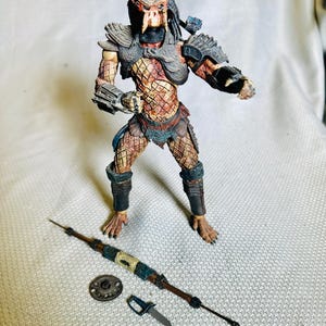 May include: A detailed action figure of a Predator alien, standing with a spear, a small shield, and a knife. The figure is predominantly brown, with intricate armor and a detailed head. The accessories are also brown and metallic.