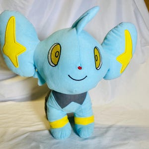 Peut inclure: Peluche d'une créature bleue avec de grandes oreilles, des éclairs jaunes et des yeux jaunes. Le jouet a une poitrine grise et des bandes jaunes sur les jambes. Il a un petit nez rouge et une bouche souriante.