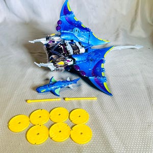 May include: A blue and purple toy spaceship with white accents, a blue toy shark, two yellow rods, and several yellow circular pieces. The spaceship has a detailed design and is set against a neutral background.