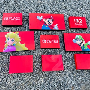 Könnte beinhalten: Eine Sammlung roter Nintendo Switch Konsolenhüllen mit den Charakteren Mario, Luigi und Prinzessin Peach. Einige Hüllen zeigen das Nintendo Switch Logo, andere die Zahl 2.