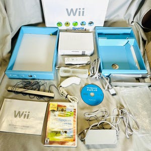 Puede incluir: Un conjunto completo de consola de juegos Nintendo Wii blanca, que incluye la consola, los mandos, el disco del juego y los manuales. La caja es blanca con el logotipo de Wii y gráficos de Wii Sports. El conjunto se muestra sobre una superficie neutra.