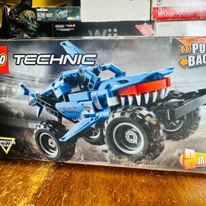 Puede incluir: Una caja de set de construcción LEGO Technic Monster Jam Megalodon con un camión monstruo azul con forma de tiburón, neumáticos negros grandes, dientes blancos y motor de retroceso. El empaque muestra el número 42134, 7+ y un logo de 2 en 1.