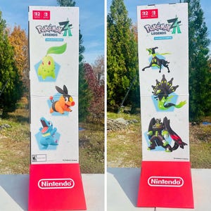 Könnte beinhalten: Ein hoher, rechteckiger Aufsteller mit dem Spiel "Pokémon Legends Z". Der Aufsteller hat einen roten Sockel mit dem Nintendo-Logo. Die linke Seite zeigt drei Pokémon-Charaktere, während die rechte Seite drei verschiedene Pokémon-Charaktere zeigt. Der Hintergrund ist eine natürliche Umgebung im Freien.