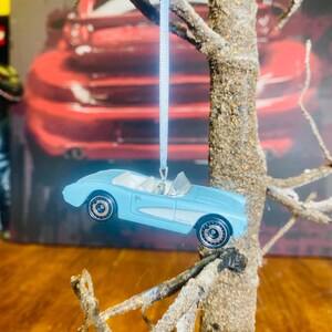 Può includere: Un ornamento per auto d'epoca blu chiaro appeso a un ramo di un albero ricoperto di neve. L'auto ha interni bianchi e ruote nere.