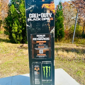Könnte beinhalten: Ein hoher, rechteckiger Werbedisplay für "Call of Duty: Black Ops 6" mit einer Soldaten-Grafik. Es enthält Text für "Bonus In-Game Rewards" und einen QR-Code. Darunter befinden sich Bilder von Monster Energy-Getränkedosen.