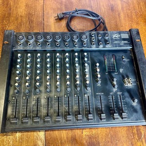 以下が含まれることがあります： 複数のノブ、スライダー、入力ジャックを備えた黒いPeavey Unity Series 1000オーディオミキサー。ミキサーには電源コードが接続されており、木製の表面にあります。