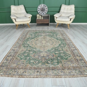 May include: A green and beige vintage rug with a floral pattern. The rug is laid out on a light wood floor in a room with two white chairs and a green wall.