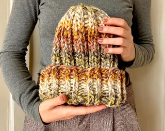 Può includere: Un berretto lavorato a maglia a mano in filato multicolore con sfumature di bianco, arancione, marrone e verde. Il berretto è tenuto da una persona che indossa un maglione a collo alto grigio e pantaloni marroni.