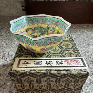 May include: A decorative, hand-painted porcelain bowl with a yellow interior and a green rim. The bowl features a dragon design and floral accents. It sits atop a patterned box with Chinese characters.