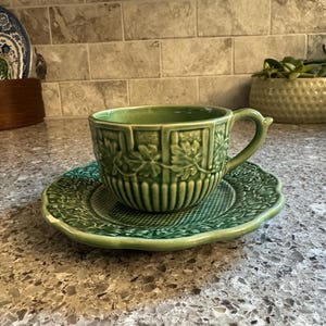 May include: A sage green ceramic teacup and saucer set featuring a raised relief pattern of grapevines and leaves. The cup has a vertical ribbed design around the base, while the saucer has a scalloped edge and a textured grid pattern in the center.