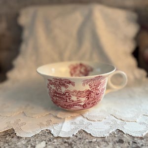 May include: A white teacup with a red and white pastoral scene design. The teacup has a handle and sits on a white, scalloped-edge doily. The cup's design features buildings, trees, and figures.