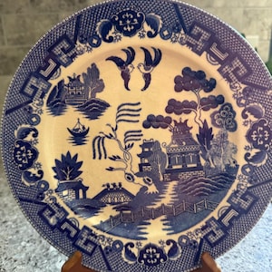 May include: A round ceramic plate featuring a blue and white willow pattern. The design depicts a landscape with a pagoda, a bridge, a boat on water, and two birds flying at the top. The rim has a geometric border with floral accents.