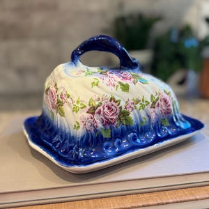 May include: A ceramic butter dish with a domed lid featuring a floral pattern of pink roses and green leaves. The base and the top handle of the lid are finished in a deep cobalt blue glaze with a scalloped, embossed edge design along the bottom.