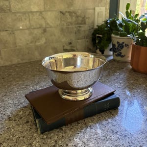 May include: A polished silver-toned bowl with a smooth, reflective surface and a circular pedestal base. The bowl is resting on top of a brown hardcover book, which is positioned on a dark blue book with visible text on the spine.
