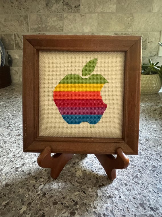 Hand-stitched cross stitch recreation of the iconic rainbow Apple logo in a tiny wooden frame — a perfect pixel-art-before-pixels artifact of computing history.