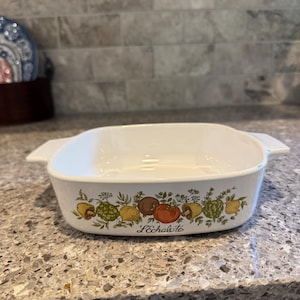 May include: A white square ceramic baking dish with integrated side handles. The exterior features a colorful botanical illustration of various vegetables including tomatoes, mushrooms, and herbs, with the text L'Echalote printed in black script.