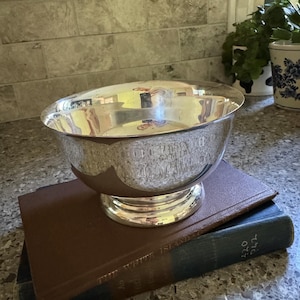 May include: A polished silver-toned trophy bowl with a wide rim and a circular pedestal base. The exterior of the bowl features an engraved inscription that reads, "COURTESY CUP GYM DIV. CLUB CHAMPIONSHIP 1980." The bowl is resting on two stacked hardcover books.