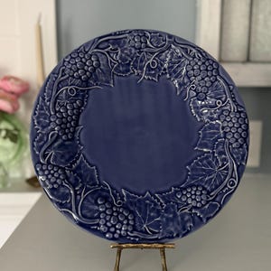 May include: A round, navy blue ceramic plate with a raised grape and leaf design around the rim. The plate is displayed on a small, gold-colored metal stand. The center of the plate is a solid, smooth navy blue.