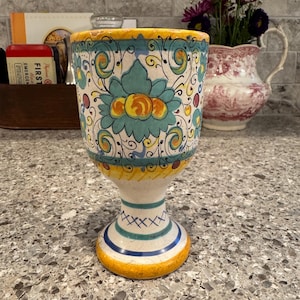 May include: A hand-painted ceramic goblet with a floral design in teal, yellow, and orange. The goblet has a wide rim and a pedestal base with blue and yellow stripes. The background includes a gray countertop and a decorative pitcher.