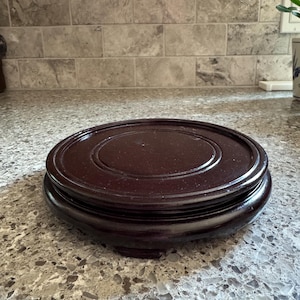 May include: A round, dark brown wooden display stand with a smooth, polished finish. The stand features a raised circular platform with concentric grooved rings on the top surface and is supported by short, sturdy feet at the base.