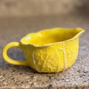 May include: A bright yellow ceramic creamer or small pitcher featuring a textured surface that mimics the appearance of cabbage leaves. The vessel has a curved handle on one side, a scalloped rim, and a glossy finish that highlights the veins.