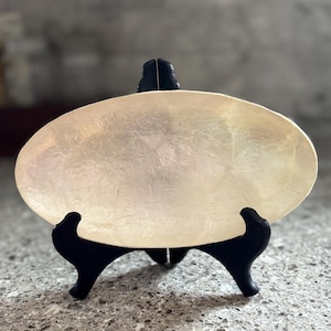 May include: An oval, iridescent serving dish with a pearlescent finish. The dish is displayed on a black, decorative stand. The background is a blurred, neutral color.
