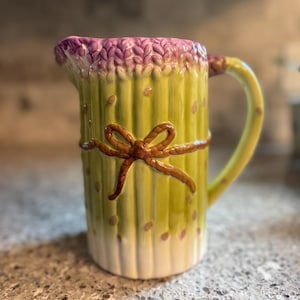 May include: A ceramic pitcher shaped like a bundle of asparagus stalks. The vessel features a light green body with a white base, a purple-tipped rim, and a brown sculpted bow tied around the center. It includes a curved handle on the side.