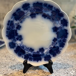 May include: A white and blue antique plate with a scalloped edge. The plate features a blue floral design around the rim and a white center. The plate is displayed on a black stand.
