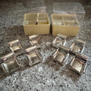 May include: A set of eight silver-toned metal miniature armchair place card holders. Each piece is shaped like a small club chair with a smooth, reflective finish. Two tan cardboard boxes with clear plastic lids are included in the set.