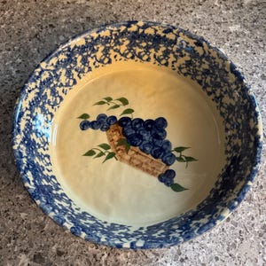 May include: Round ceramic serving dish with a cream-colored interior and a blue speckled rim. The center features a cluster of blue grapes in a brown basket with green leaves. A decorative piece for the kitchen.