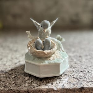 May include: A porcelain figurine of a bird with outstretched wings, perched above a nest with two baby birds. The nest sits atop a hexagonal, light blue and white base. The figurine is a soft, muted color.