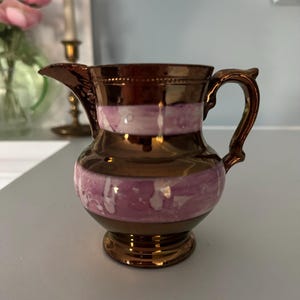May include: A small, antique creamer with a bronze finish and a decorative handle. The creamer features a band of pink and white paint around the middle. The creamer has a spout for easy pouring and sits on a small base.