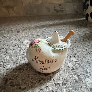 May include: A ceramic mustard pot with a cream-colored glaze, featuring a hand-painted pink rose design and the black cursive text "Moutarde fine". It includes a matching lid with a small handle and a wooden spoon inserted into the opening.