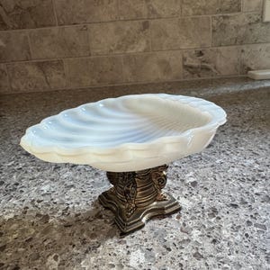 May include: A white, translucent, shell-shaped dish with a scalloped edge, supported by a dark, metallic, pedestal-style base featuring ornate, sculpted details. The bowl has a smooth, curved interior with subtle ridges radiating from the center.