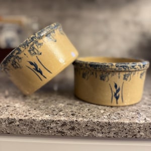 May include: Two beige ceramic bowls with blue floral patterns. The bowls have a slightly flared rim and a decorative blue border. The bowls are decorated with blue wheat-like sprigs. The bowls are sitting on a speckled countertop.