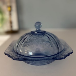 May include: A blue glass butter dish with a domed lid and a decorative floral pattern. The dish has a round knob on top of the lid and a scalloped edge. The butter dish is made of glass and is a vintage item.