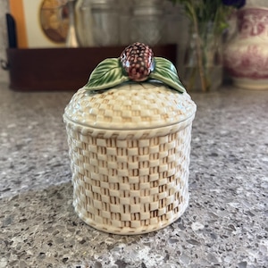 May include: A ceramic jar with a woven basket design. The lid is topped with a detailed blackberry and green leaves. The jar is off-white with brown accents, and sits on a speckled countertop. A decorative piece for the kitchen.