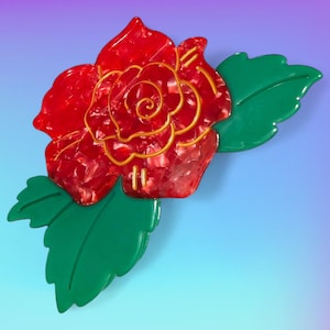 May include: A red rose brooch with gold accents and green leaves. The rose has a textured appearance, and the leaves are a solid green color. The brooch is set against a gradient background of blue and purple.